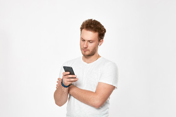 Studio shot of a bearded young man with wireless headphones holding looking at the screen of a mobile phone.