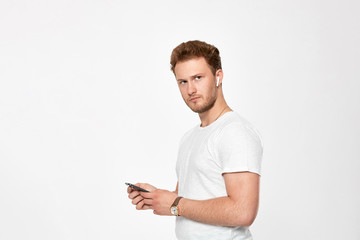 Studio shot of a bearded young man with wireless headphones holding mobile phone and looking at the camera.