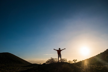 teenager in mountain stay with open arms alone and with headphones and a great sunset - jogging  and fit - sun at the background and sky - freedom concept - caucasian 20s