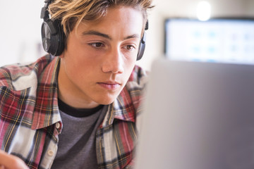 caucasian teenager indoor doing homework on the table at home - blonde guy writing and reading in his laptop or computer to get greats scores - portrait of boy with headphones