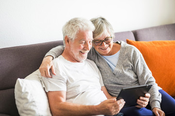 couple of seniors smiling and looking at the same tablet hugged on the sofa - indoor, at home concept - caucasians mature and retired man and woman using technology