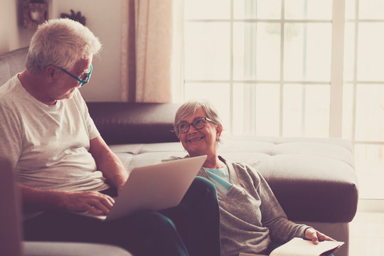 couple of seniors at home - man with laptop and glasses sitting at the sofa - woman sitting on the grund reading a book - indoor and love concept
