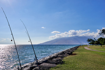 A view of fishing rod anchored between rocks,  Hawaiian ocean, and an island seen from a park in Maui.