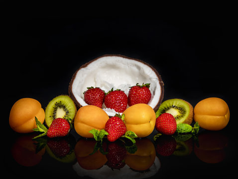 Fruit platter of apricots, kiwi, half coconut, strawberries and mint, on a transparent dark background with reflection.