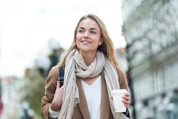 Pretty young woman holding cup of coffee while walking in the street.