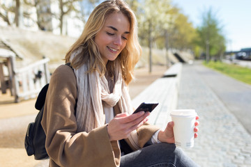 Pretty young woman using her mobile phone while holding cup of coffee in the street.