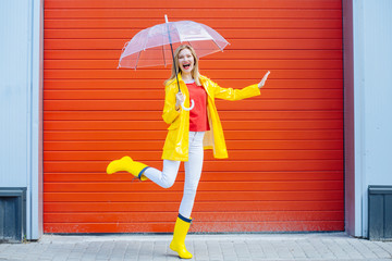 Emotional woman dancing laughing under umbrella in yellow raincoat, rubber boots, red t-shirt over red garage door background. Cool expression on face. Outdoor. Full height. Weather forecast concept