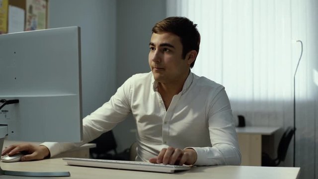 Young handsome businessman in white stylish shirt sits down at the table, checks the phone and starts working at the desktop computer in the office. Trader looking at screen monitoring stock online