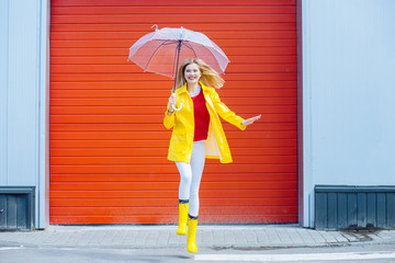 Emotional woman dancing laughing under umbrella in yellow raincoat, rubber boots, red t-shirt over red garage door background. Cool expression on face. Outdoor. Full height. Weather forecast concept
