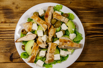 Tasty salad with fried chicken breast, green olives, feta cheese, avocado, lettuce and olive oil on wooden table. Top view