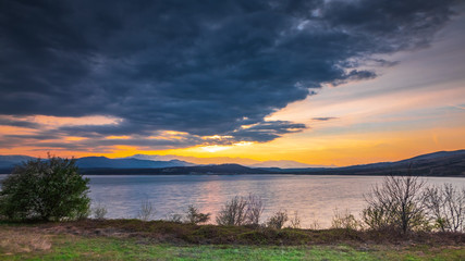 A sunset on dark sky with dramatic clouds over a lake with distant hills in the background and a few trees and bushes in the foreground