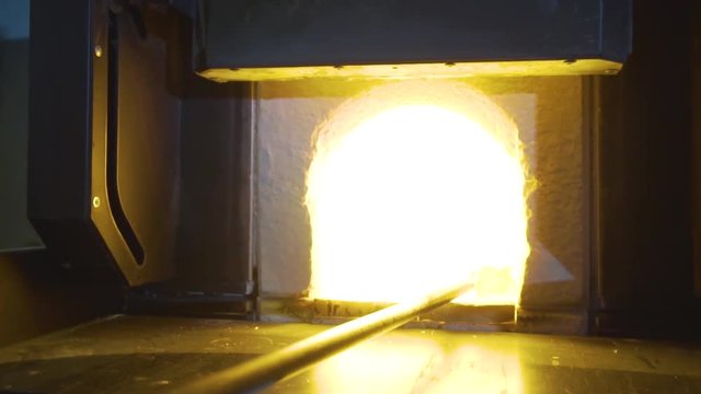 Glassblower in his workshop taking the rod with liquid glass out of glowing hot oven.