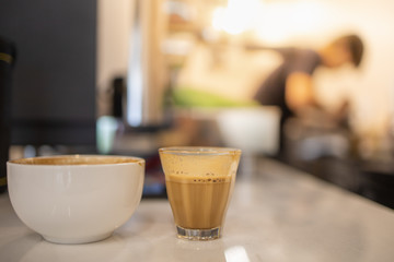 Close up of shot glass and white cup of hot coffee latte with coffee maker machine and barista as background.