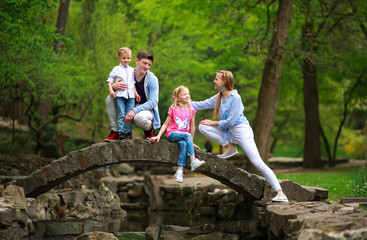Naklejka na ściany i meble Happy young family with children in summer green park on stone bridge over the river in forest