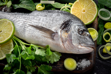 Fresh and raw dorada fish with lemon, onion and herbs on a black wooden background. preparation in the kitchen