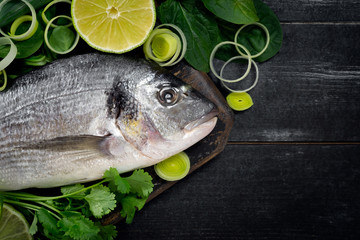 Fresh and raw dorada fish with lemon and herbs on a black wooden background, top view and copy space