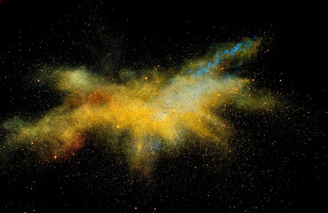 Bizarre forms of golden glitter powder paint explode in front of a black background to give off...