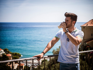 Handsome man drinking american coffee from big paper cup, outdoor standing by handrail in Italian sea town in a summer day. Horizontal outdoors shot