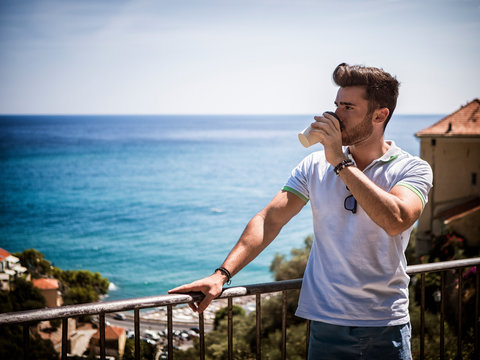 Handsome Man Drinking American Coffee From Big Paper Cup, Outdoor Standing By Handrail In Italian Sea Town In A Summer Day. Horizontal Outdoors Shot