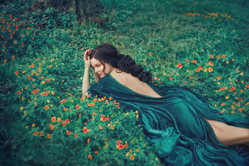 charming nymph lying on green grass and bright small flowers, girl in a long emerald velor dress...