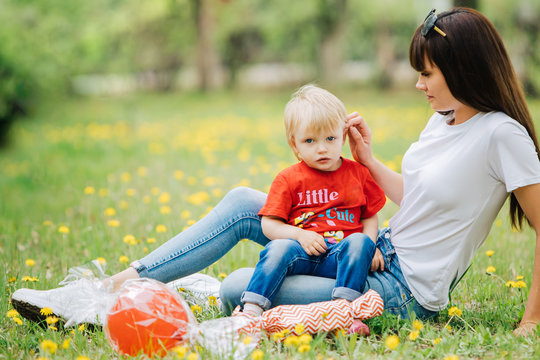 A beautiful young mother sits with her blond baby on in the green grass and dandelions. Photo in the park on the background of green foliage.
