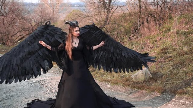 dark angel with large massive black wings and horns on head stands in middle of road in long flying vintage dress with lace sleeves. girl listens to singing of wind, lady poses for fantastic photos