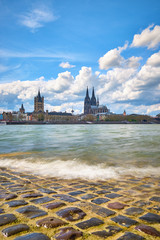 Stadtportrait Kölner Dom, Groß St Martins Kirche mit Blick über den Rhein und dem Ufer im Vordergrund mit einer wolkigen Welle und römischen Asphalt.