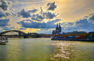 Sonnenuntergang in Köln mit Blick auf Rhein Hohenzollermbrücke Musicla Dome und ein goldenes Wolkengebilde mit Blauen goldenen Farben. Auf dem Rhein zieht ein Schiff vorbei.