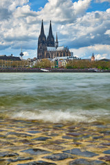 Stadtportrait Kölner Dom, Groß St Martins Kirche mit Blick über den Rhein und dem Ufer im Vordergrund mit einer wolkigen Welle und römischen Asphalt.