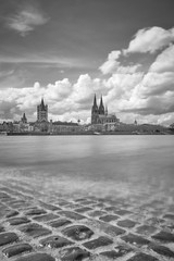 Schwarz Weiß portrait vom Kölner Dom und der Groß St. Martins Kirche mit Blick auf den Rhein und altes römisches Ufer im Vordergrund. Über den Himmel schweben schöne fluffige dichte Wolken.