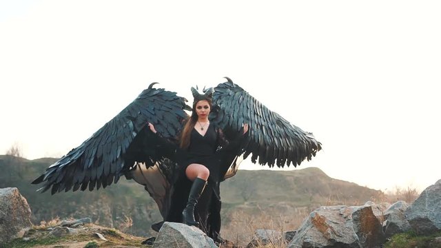 graceful lady with a menacing look looks into the soul, a girl in a long black dress with lace sleeves lifts the hem of her dress and her heavy strong feather wings, a dark angel fallen from heaven