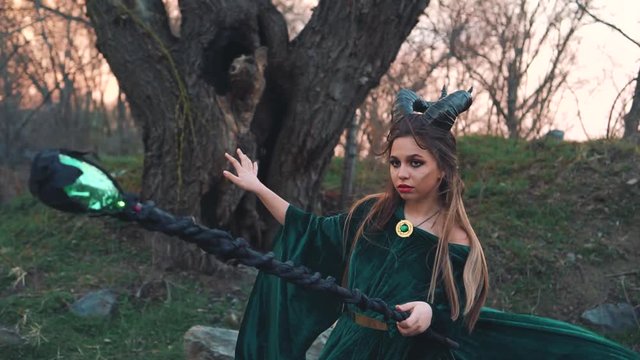 strict witch with magical magical glowing and sparkling staff in her hands, lady with horns on head utters spell, emerald silk flying long dress, demonessa on grassy bank of river, magic spell