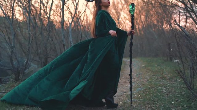 dark witch stands on forest path in long flying emerald dress free flying sleeves with a golden belt. lady with magic stick in hand collects energy of forest and the sun for evil, creative art