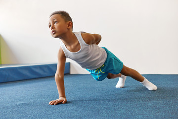 Disciplined skinny African American kid in sportswear doing single arm plank, keeping one hand on lower back, trying to hold it, looking up. Fitness, active lifestyle and childhood concept