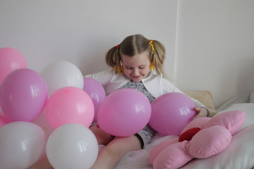 Fototapeta na wymiar a girl plays in bed with white and pink balloons and a strawberry pillow