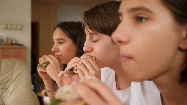 Triplets sisters teenager girls at home watch tv sit on a couch eating tasty sandwiches together