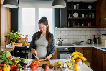 Beautiful smiling young pregnant woman preparing healthy food with lots of fruit and vegetables at home kitchen