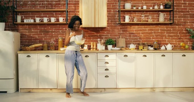 Charming African American happy woman in pajama having fun and dancing in the kitchen early in the morning before breakfast.