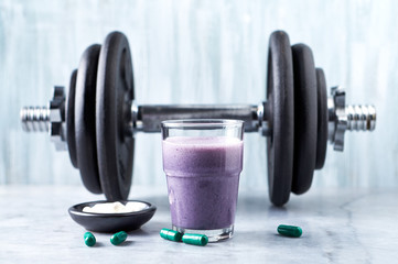 Fototapeta na wymiar Glass of Protein Shake with milk and blueberries, Beta-alanine and L-Carnitine capsules and a dumbbell in background. Sports bodybuilding nutrition. Stone / Wooden background. Copy space. 