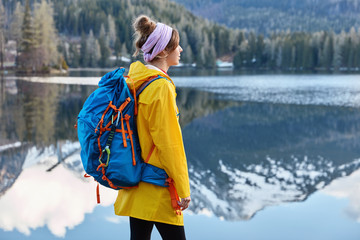 Outdoor view of thoughtful restful woman stands near calm lake with mountain reflection, has tourist backpack, wears yellow raincoat, stands against turquoise water at shore. Relax time on holiday