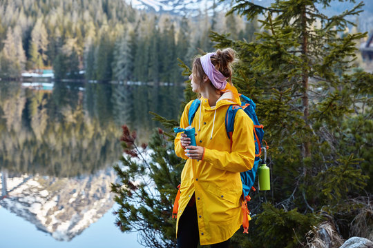 Horizontal outdoor view of calm female tourist admires beautiful mountains, forest and lake, keeps gaze aside, stands alone, wears yellow raincoat. People, lifestyle and active vacation concept
