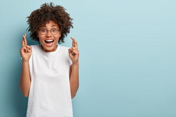 Studio shot of happy dark skinned woman crosses fingers for good luck, wears round glasses and...