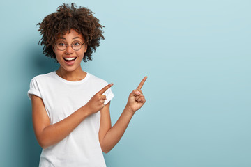Happy smiling woman seller points at upper right corner, advertises item on blank space, has friendly facial expression, wears casual white t shirt, isolated on blue background. Look there, its nice