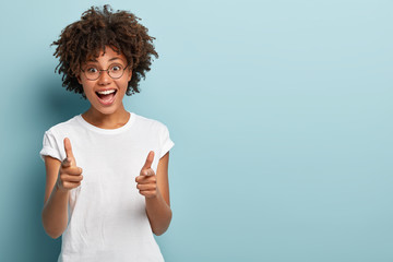 Joyous dark skinned woman points both fore fingers straightly at camera, wears casual clothing, happy to choose someone, isolated over blue background with empty space for your text or promotion
