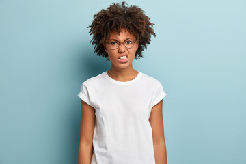 Pissed annoyed woman has Afro hairstyle, clenches teeth from anger, stares with hate, frowns face, wears round specacles and white casual t shirt, models on blue background, expresses hateful emotions