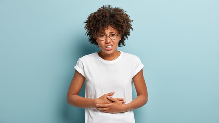 Dissatisfied young woman has food poisoning, keeps hands on belly, suffers from menstrual period cramp, feels discomfort in stomach, wears casual clothes, frowns face, isolated on blue background - obrazy, fototapety, plakaty