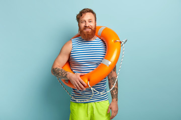 Happy redhaired male vacationist wears sailor vest and short, carries orange lifebuoy for save...