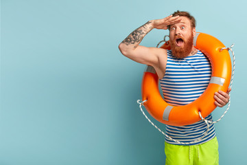 Surprised young holiday maker with red hair and beard, comes on beach with safety equipment as...