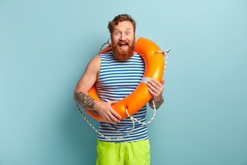 Overjoyed male lifeguard with tattoo, foxy beard, poses with inflated rescue ring, prevents...