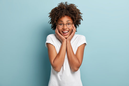 Optimistic carefree Afro female touches both cheeks, has broad smile, shows white teeth, dressed in casual outfit, round spectacles, stands against blue background, grins at camera, enjoys life